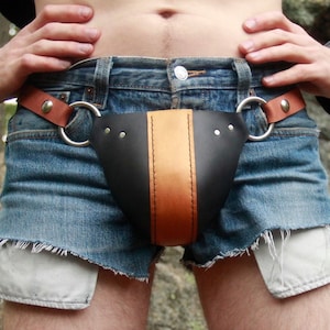 Könnte beinhalten: Ein schwarzer Ledergurt mit orangefarbenem Streifen, getragen über einer Jeans-Shorts. Der Gurt hat zwei silberne D-Ringe und einen braunen Lederriemen.