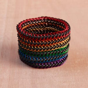 Pode incluir: Uma pilha de pulseiras circulares de malha de corrente feitas de an&eacute;is de metal entrela&ccedil;ados. Cada pulseira apresenta uma cor distinta, incluindo vermelho, laranja, dourado, verde, azul e roxo, combinadas com an&eacute;is pretos.
