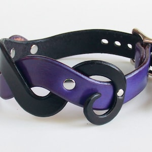 May include: A purple leather bracelet with a black leather accent in the shape of an infinity symbol. The bracelet has a silver buckle.