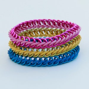Pode incluir: Uma pilha de tr&ecirc;s pulseiras de malha met&aacute;lica com an&eacute;is de metal entrela&ccedil;ados. A pulseira superior &eacute; rosa brilhante, a pulseira do meio &eacute; dourada e a pulseira inferior &eacute; azul. Cada pulseira &eacute; constru&iacute;da com um padr&atilde;o de tecelagem consistente.