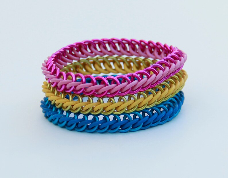 Pode incluir: Uma pilha de tr&ecirc;s pulseiras de malha met&aacute;lica com an&eacute;is de metal entrela&ccedil;ados. A pulseira superior &eacute; rosa brilhante, a pulseira do meio &eacute; dourada e a pulseira inferior &eacute; azul. Cada pulseira &eacute; constru&iacute;da com um padr&atilde;o de tecelagem consistente.