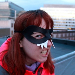 May include: A woman with red hair wearing a black leather mask with white teeth. The mask covers her eyes and part of her forehead. She is wearing a red jacket and has a serious expression on her face.