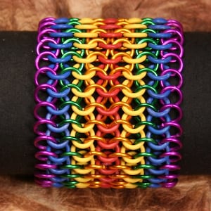 Pode incluir: Uma pulseira larga e colorida feita de anéis de metal interligados. A pulseira apresenta um arco-íris de cores, incluindo roxo, azul, verde, amarelo, laranja e vermelho. A pulseira é exibida em uma superfície preta.