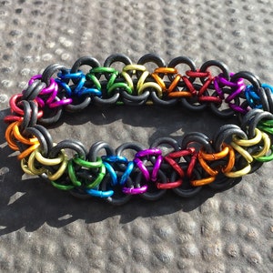 Pode incluir: Um bracelete de malha de corrente arco-íris com anéis pretos. O bracelete é feito de pequenos anéis de metal que são ligados uns aos outros em um padrão.