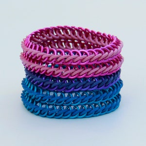Pode incluir: Uma pilha de quatro pulseiras de malha met&aacute;lica com an&eacute;is de metal entrela&ccedil;ados num gradiente de cores. As duas pulseiras superiores s&atilde;o em tons de rosa e magenta, enquanto as duas inferiores s&atilde;o em tons de roxo escuro e azul.