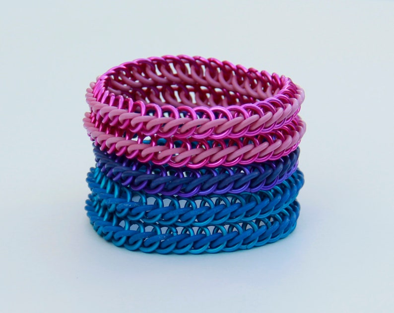 Pode incluir: Uma pilha de quatro pulseiras de malha met&aacute;lica com an&eacute;is de metal entrela&ccedil;ados num gradiente de cores. As duas pulseiras superiores s&atilde;o em tons de rosa e magenta, enquanto as duas inferiores s&atilde;o em tons de roxo escuro e azul.