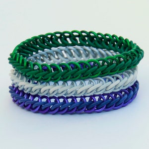 Pode incluir: Uma pilha de tr&ecirc;s pulseiras de malha met&aacute;lica com an&eacute;is entrela&ccedil;ados. A pulseira superior &eacute; verde vibrante, a do meio &eacute; cinza claro fosco e a inferior &eacute; de um roxo profundo e saturado. Cada pulseira &eacute; constru&iacute;da com um padr&atilde;o consistente e repetitivo de elos circulares.