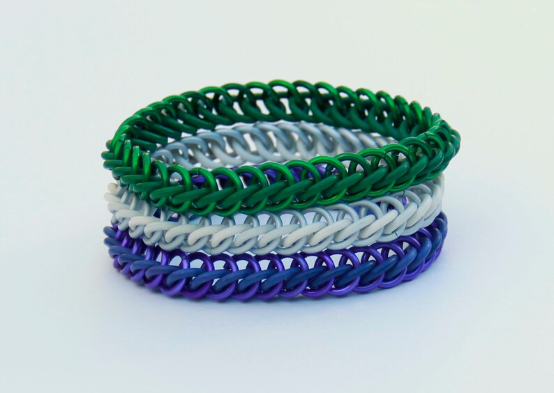Pode incluir: Uma pilha de tr&ecirc;s pulseiras de malha met&aacute;lica com an&eacute;is entrela&ccedil;ados. A pulseira superior &eacute; verde vibrante, a do meio &eacute; cinza claro fosco e a inferior &eacute; de um roxo profundo e saturado. Cada pulseira &eacute; constru&iacute;da com um padr&atilde;o consistente e repetitivo de elos circulares.