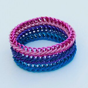 Pode incluir: Uma pulseira de malha de metal constru&iacute;da com an&eacute;is met&aacute;licos entrela&ccedil;ados num gradiente de cores, apresentando uma camada superior rosa brilhante, uma camada m&eacute;dia roxo escuro e uma camada inferior azul vibrante, criando um visual empilhado.