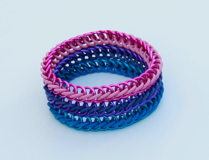 Pode incluir: Uma pulseira de malha de metal constru&iacute;da com an&eacute;is met&aacute;licos entrela&ccedil;ados num gradiente de cores, apresentando uma camada superior rosa brilhante, uma camada m&eacute;dia roxo escuro e uma camada inferior azul vibrante, criando um visual empilhado.