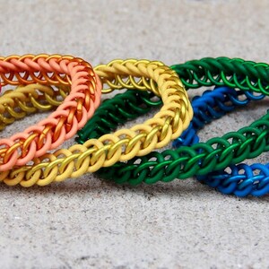 Pode incluir: Um conjunto de seis pulseiras circulares de cota de malha dispostas em fila, apresentando um espectro vibrante de cores, incluindo vermelho, laranja, amarelo, verde, azul e roxo. Cada pulseira &eacute; feita de an&eacute;is de metal entrela&ccedil;ados em um padr&atilde;o uniforme.