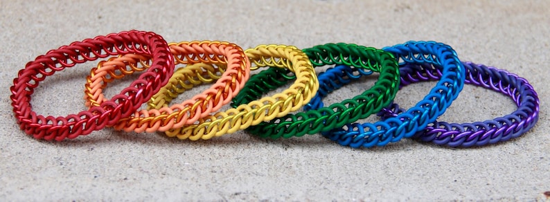 Pode incluir: Um conjunto de seis pulseiras circulares de cota de malha dispostas em fila, apresentando um espectro vibrante de cores, incluindo vermelho, laranja, amarelo, verde, azul e roxo. Cada pulseira &eacute; feita de an&eacute;is de metal entrela&ccedil;ados em um padr&atilde;o uniforme.