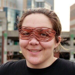 May include: A person wearing a brown leather mask with a cut-out design. The mask covers the eyes and part of the nose. The person is smiling.