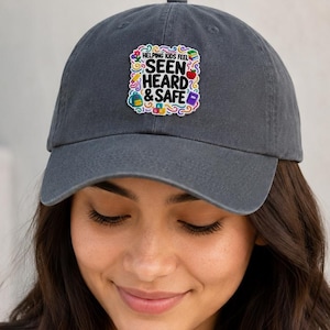 May include: A charcoal gray baseball cap featuring a square patch on the front. The patch has a colorful, swirling border and the text "HELPING KIDS FEEL SEEN HEARD & SAFE" in black, accompanied by small icons of a backpack, apple, and book.