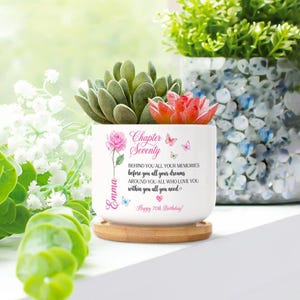 Puede incluir: Una maceta de cerámica blanca con base de madera, con una planta suculenta y una flor roja. La maceta tiene el texto "Chapter Seventy" y un nombre personalizado. El texto dice "¡Feliz 70 cumpleaños!"