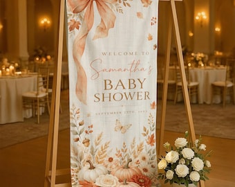 Peut inclure: Une bannière en tissu verticale sur un support en bois avec un grand nœud pêche en haut, des feuilles d'automne, des citrouilles et un papillon. Le texte indique : Welcome to Samantha's Baby Shower, September 25th, 202X. Un bouquet floral blanc est posé à côté.
