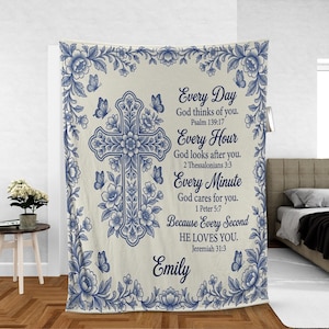 May include: A cream-colored blanket with a blue floral and butterfly design. A large cross is centered, surrounded by flowers. Text reads "Every Day God thinks of you" and other inspirational phrases. The name "Emily" is at the bottom.