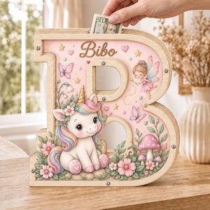 May include: A wooden letter B shaped money box with a light pink background featuring a cartoon unicorn, a fairy, butterflies, flowers, and mushrooms. The name Bibo is printed at the top. The frame is secured with visible metal screws.