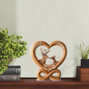 May include: A wooden heart-shaped decorative piece featuring two deer kissing inside. The names "Edward" and "Selena" are inscribed, with the phrase "You & Me We Got This" below. The piece has a light brown finish and an infinity base.