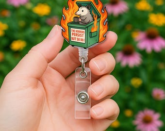 May include: A retractable badge reel featuring a cartoon opossum in a burning green trash can, holding a mug. The reel has the text "THE HORRORS PERSIST BUT SO DO I". The design is surrounded by orange flames, with a clear plastic strap and a metal button.
