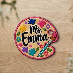 May include: A circular embroidered patch shaped like an artist's palette with a tan background and a pink border. The text Ms. Emma is embroidered in black. The design features colorful paint splatters, small flowers, and a paintbrush.