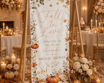 May include: A vertical fabric banner on a wooden stand featuring the text Fall In Love WELCOME TO Amanda's BRIDAL SHOWER. The design includes autumn leaves, pumpkins, acorns, and two champagne flutes with a white wood-grain background.