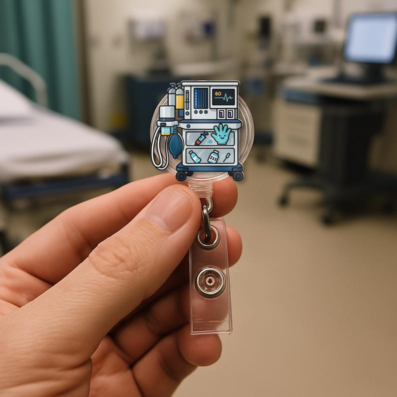 May include: A clear, round badge reel featuring a cartoon illustration of a medical ventilator on a rolling cart. The ventilator has a screen displaying a waveform. The reel is held by a hand in a medical setting.