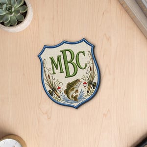 May include: A shield-shaped embroidered patch featuring the green monogram letters MBC at the top. Below the letters is a detailed brown fish jumping out of blue water, surrounded by fishing rods, reels, and red and white bobbers.
