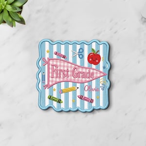 May include: A square embroidered patch with a scalloped edge featuring a light blue and white striped background. A pink gingham pennant displays the text First Grade. The design includes a red apple, scissors, and colorful crayons.