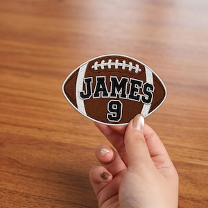 May include: An embroidered patch shaped like an American football, featuring a brown background with white laces and stripes. The text JAMES is stitched in black with a white outline, and the number 9 is centered below in the same style.