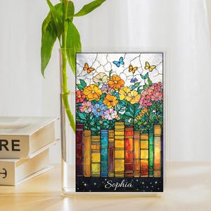 Könnte beinhalten: Eine farbenfrohe Glasmalerei mit einem lebendigen Blumenarrangement mit Schmetterlingen und einer Reihe von Büchern. Das Panel enthält den Namen "Sophia" am unteren Rand. Das Kunstwerk ist in einem transparenten Rahmen.
