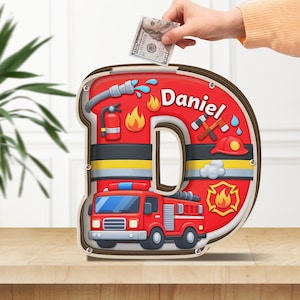 May include: A wooden letter "D" shaped piggy bank with a firefighter theme. The bank is red with yellow and black stripes, and features a fire truck, flames, and the name "Daniel". A hand is inserting a $100 bill.