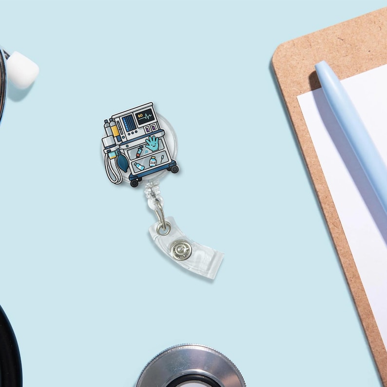 May include: A badge reel featuring a medical cart design with a monitor, gauges, and medical supplies. The reel has a clear plastic clip and a retractable cord. The background is a light blue surface with a stethoscope, clipboard, and pen.