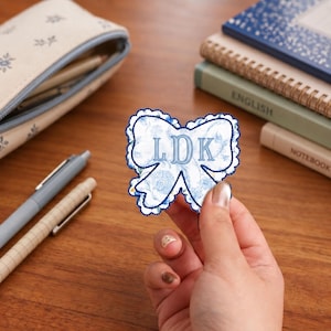 May include: A bow-shaped fabric patch featuring a blue and white floral pattern with a scalloped dark blue edge. The center of the patch is embroidered with the letters LDK in a light blue serif font, creating a decorative monogram design.