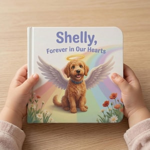 Può includere: Un libro per bambini intitolato "Shelly, Forever in Our Hearts" è tenuto da una persona. La copertina presenta un cane dei cartoni animati con ali d'angelo e un alone, su uno sfondo arcobaleno. Illustrazioni floreali in basso.