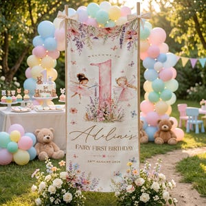 Può includere: Uno striscione verticale con un grande numero uno rosa, due illustrazioni di fate e decorazioni floreali. Il testo recita Welcome to Adeline's Fairy First Birthday 28th August 2026. Lo striscione è esposto all'aperto con archi di palloncini.