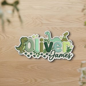 May include: A custom embroidered patch featuring the name Oliver James in various shades of green. The design includes a T-Rex, a long-necked dinosaur, a volcano, and small stars, all outlined in white with a textured fabric background.