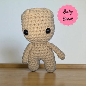 May include: A crocheted Baby Groot doll in a light tan color. The doll has large black eyes and a small body with arms and legs. The text "Baby Groot" is in a pink starburst shape in the upper right corner. The doll is on a wooden surface.