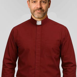 May include: A burgundy dress shirt with a white clerical collar and a chest pocket. The shirt has long sleeves and is buttoned up. The person in the image is wearing a black belt and dark pants.