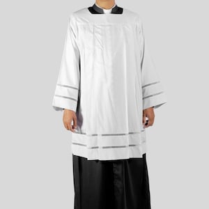 Op de afbeelding: Een witte liturgische albe met decoratieve horizontale banden op de mouwen en zoom. De albe wordt gedragen met een zwarte soutane. De persoon op de afbeelding draagt een bril en een witte priesterboord.