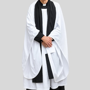 Op de afbeelding: Een persoon in een witte klerikale jurk met een zwarte stola en manchetten. De persoon draagt een witte klerikale kraag en een zwarte broek. De achtergrond is grijs.