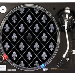 May include: A black turntable with a silver platter and a silver tone arm. The platter has a black and white design with a fleur-de-lis pattern. The turntable is labeled "Technics Quartz Direct Drive Turntable System SL-1200MK5".