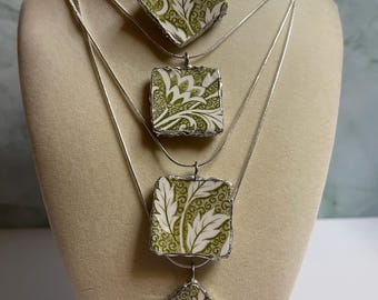 Può includere: Quattro collane con ciondoli quadrati e a forma di diamante. I ciondoli presentano un motivo floreale bianco e verde, incastonati in metallo color argento. Le collane sono esposte su un manichino beige.
