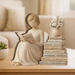 May include: A wooden figurine of a woman reading a book labeled "SOPHIA." Beside her is a stack of books with religious quotes and a small vase of flowers. The figurine is light brown, and the books are beige.
