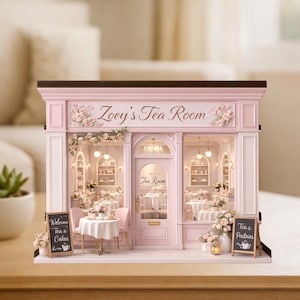 May include: A miniature pink tea room model with the text "Zoey's Tea Room" above the entrance. The model features tables, chairs, and decorative floral arrangements. Two small chalkboards display "Welcome Tea & Cakes" and "Tea & Pastries".