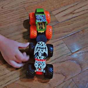 May include: Two toy monster trucks are positioned in a line. The top truck is green with bright orange wheels and features the text Wild Trash on the roof. The bottom truck is white with black spots, resembling a dalmatian, and has black wheels.