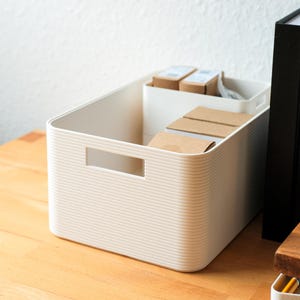May include: A cream-colored, rectangular storage bin with a ribbed texture and a rectangular handle cut-out. Inside, there's a white container holding small cardboard boxes. The bin sits on a wooden surface.