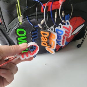 May include: A collection of colorful name tags with various names, including "Mom", "Dad", and "Ben". The tags are attached to a backpack with a Spider-Man design. The tags are made of plastic and have a 3D appearance.