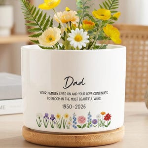 May include: A white cylindrical ceramic planter with a bamboo base, filled with yellow and white artificial flowers. The planter features the text "Dad", "YOUR MEMORY LIVES ON AND YOUR LOVE CONTINUES TO BLOOM IN THE MOST BEAUTIFUL WAYS", "1950-2026", and a colorful floral border.