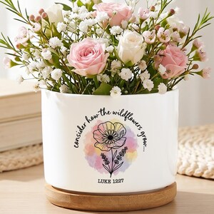 May include: A white cylindrical ceramic planter with a wooden base, filled with pink and white roses and baby's breath. The planter features a watercolour floral graphic with the text "consider how the wildflowers grow..." and "LUKE 12:27".
